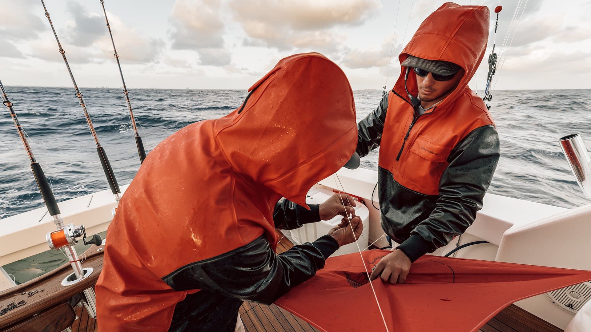 Two mates in orange foul-weather gear rig a fishing kite in the cockpit of a sportfishing boat on a rough offshore day, rods in the rocket launchers behind them.